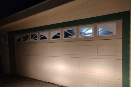 Closed white garage door with decorative window panels at the top, mounted on a house with light-colored siding and green trim.