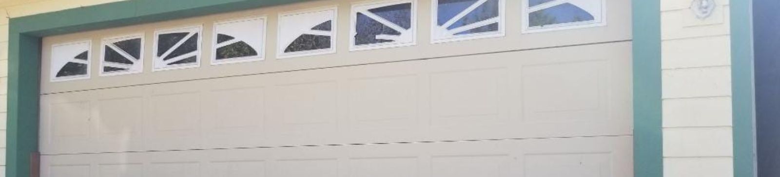 Closed white garage door with decorative window panels at the top, mounted on a house with light-colored siding and green trim.